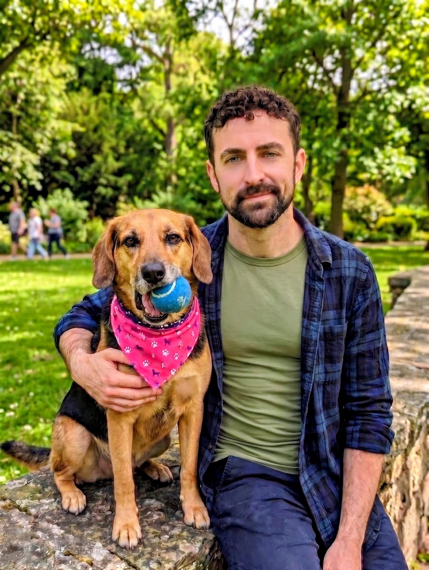 Brandon J. Redmond sitting on a low stone wall in a park with his dog, who wears a pink bandana and holds a tennis ball